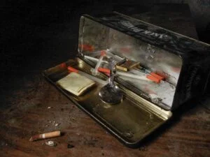 An open tin laying on its side on a wooden desk, revealing the drug paraphernalia inside and a single cigarette. The tin includes a syringes and a spoon.